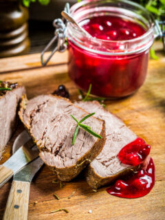 sliced instant pot venison roast on a cutting board with two knives and a jar of cranberry sauce