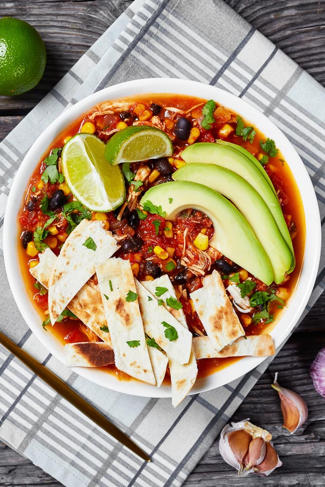 a closeup of a bowl of instant pot taco soup topped with homemade tortilla chips, avocado slices, and fresh lime wedges
