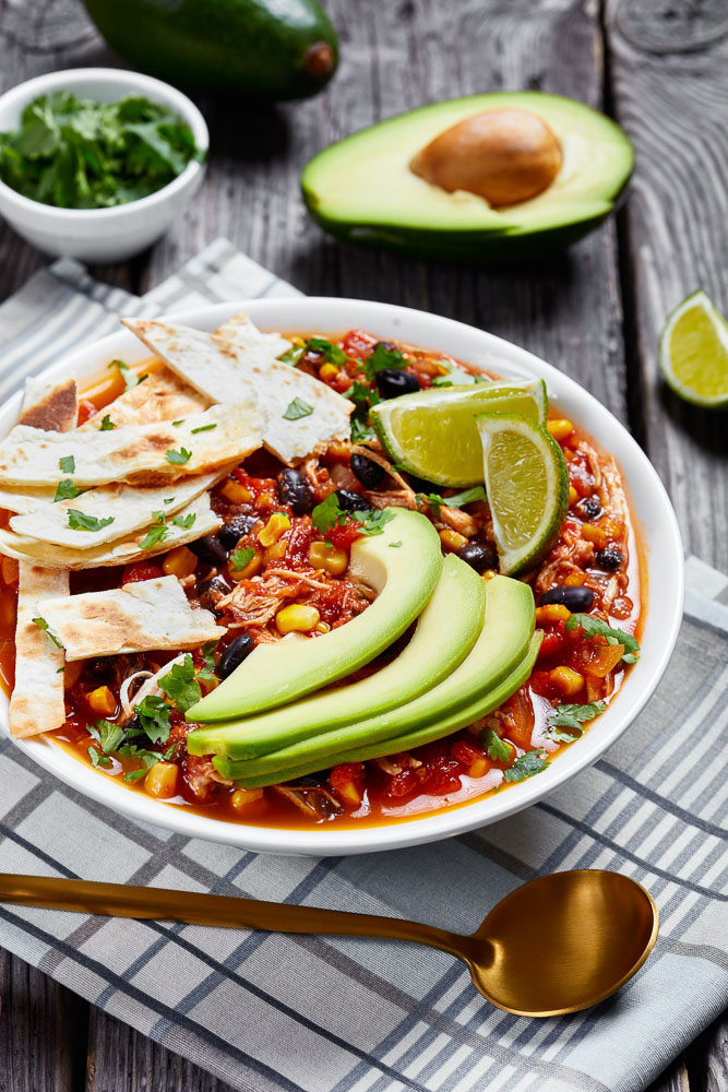 a bowl of instant pot taco soup topped with homemade tortilla chips, avocado slices, and fresh lime wedges, next to a halved avocado and a small dish of chopped cilantro